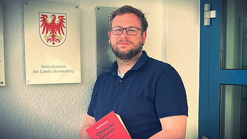 Foto zur Frage "Wie wird man Rechtspfleger*in": Zu sehen ist ein Porträt von Richard, der ein rotes Gesetzbuch in der Hand hält. Im Jobkompass stellt er die Rechtspflege vor.