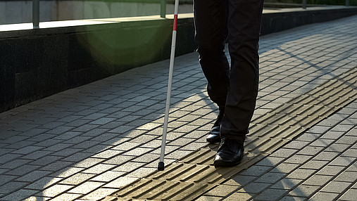 Foto zum Thema Blindheit und Leben mit einer Sehbehinderung: Zu sehen ist eine blinde Person, die sich mit einem Blindenstock an einem Leitsystem orientiert.