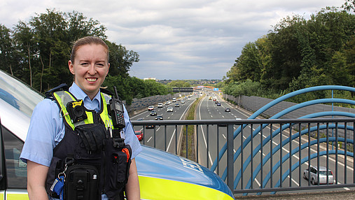 Foto zum Thema "Wie wird man Polizist*in": Zu sehen ist Svenja, die in Uniform vor dem Streifenwagen auf einer Autobahnbrücke steht.