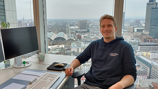 Foto zum Thema "duales Studium angewandte Informatik": Zu sehen ist Thomas, der in seinem Büro in der Bundesbank sitzt. Dieses befindet sich in einem Hochhaus, hinter ihm erstreckt sich eine beeindruckende Aussicht. 