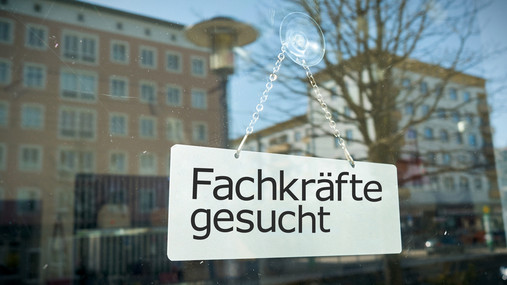 Hinter einer Fensterscheibe, in der sich Hochhäuser gegenüber spiegel, hängt ein Schild mit der Aufschrift "Fachkräfte gesucht".